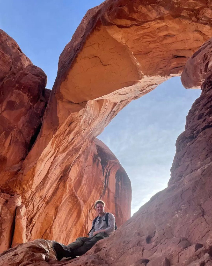 man sitting under double arch in arches national park