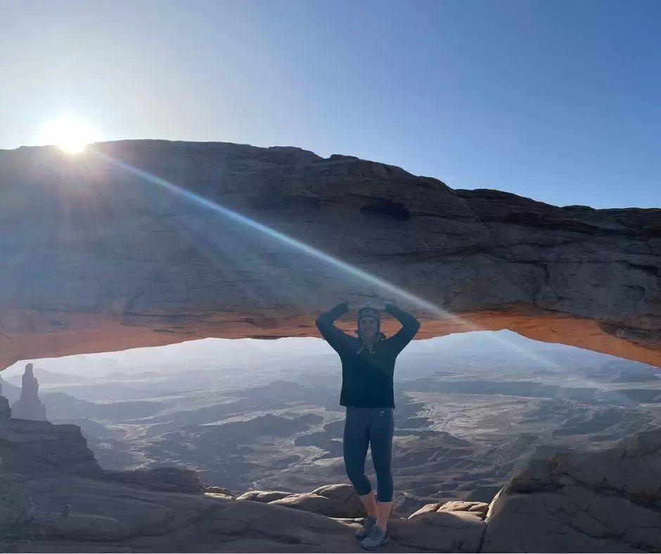woman making an arch formation with her arms while standing under mesa arch at canyonlands